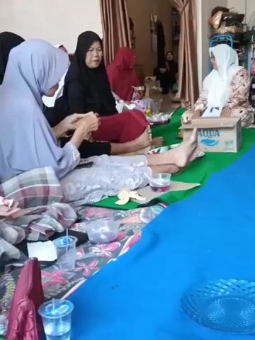 A group of people are sitting on the floor around a blue mat, eating and talking. Some are wearing hijabs. There are plates of food and drinks on the floor. A woman in a gray robe walks across the room. In the background, there is a cabinet filled with items.