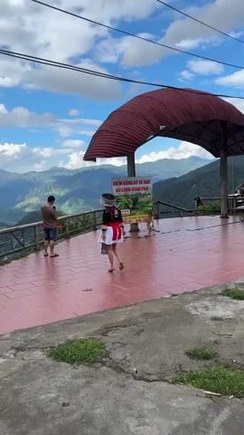 A full shot shows a woman in a colorful traditional dress walking on a tiled ground with a mountain range in the background. A man is filming on the left. A large yellow sign with Vietnamese text and pictures of a person zip-lining is visible. A red roof structure covers part of the area. A small child in a brown outfit is visible near the sign. The camera zooms in on the child, who is standing near the sign, smiling, and looking at the camera. The child then starts to walk.