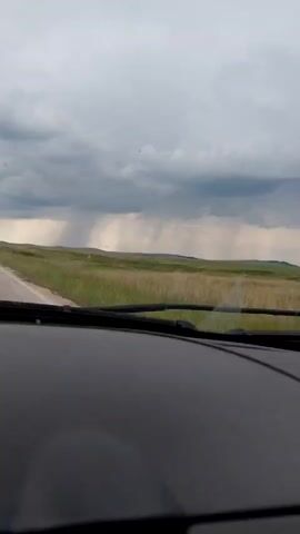 The video displays a static view from inside a vehicle, looking out through the windshield. The dark dashboard and base of the windshield wipers are visible at the bottom of the frame. A paved road stretches into the distance along the left side. The majority of the landscape consists of vast, green, grassy fields under a heavily clouded, overcast sky. In the mid-ground and distance, multiple distinct columns or sheets of rain are visible descending from the dark clouds, indicating localized precipitation. Human speech is audible in the background.