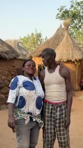 A waist-up shot shows a woman and an older man standing outside with their arms around each other. The woman is on the left, facing the camera and smiling. She has dark skin and is wearing a blue and white printed shirt, gray pants, and a printed fabric tied around her waist. The man is on the right, facing slightly to the left. He has gray hair, dark skin, and is wearing a white tank top and plaid pants. He is holding a cigarette in his right hand. Behind them are several small, mud brick houses with thatched roofs. The ground is dirt. The sky is light blue.