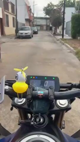 The view from the rider's seat of a motorcycle parked on a street in a residential neighborhood is seen in this short video. The motorcycle’s digital instrument panel shows the bike is in neutral and is not moving. A yellow rubber duck with a propeller on its head is attached to the motorcycle's handlebars, adding a whimsical detail to the scene. The street is narrow and lined with buildings and parked cars. There are trees and other vegetation visible on either side of the road. The sky is overcast, indicating a cloudy day. No people are seen in the video. The scene is still and quiet, providing a glimpse into a typical residential area.