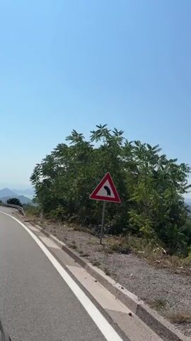 The video shows a view from a moving vehicle on a paved road. The road has white dashed lines on the left side and a white solid line on the right. The vehicle is moving along a curve. On the right side of the road, there is a metal guardrail, a concrete curb, and a dry, grassy embankment. Various trees and bushes are visible on the embankment. In the background, there are rolling hills and mountains, some of which are covered in greenery. The sky is a clear, bright blue. A few scattered white objects, possibly litter, are visible on the embankment. The overall impression is a scenic drive through a hilly, possibly coastal, landscape on a sunny day.