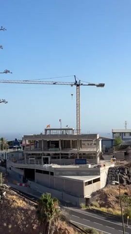 A construction site is visible under a clear blue sky. A large yellow crane is positioned over a multi-story building under construction. The building appears to be made of concrete and steel, with visible structural elements and unfinished walls. There are several flags flying from the top of the building. The site is situated on a hillside, with a road running along the bottom of the frame. Vegetation, including palm trees, is present on the surrounding slopes. The scene suggests ongoing construction activity.