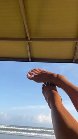 A person's feet are shown against a backdrop of the sky and ocean. The feet are bare and appear to be resting, with one foot slightly above the other. A thin ring is visible on one of the toes. The scene is framed by a yellow structure overhead. Waves are breaking on a sandy beach in the distance. The sky is blue with scattered white clouds.
