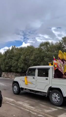 A white pickup truck with several people in the bed drives down a road. The people are wearing ceremonial robes and hats. There are flags on the truck. The truck is surrounded by trees and mountains. There are other vehicles on the road.