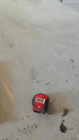 A tape measure is on the ground next to a cordless drill with a hole saw attachment. The drill is being held in a person's hand and is positioned near a wall or partition. The environment appears to be an unfinished or construction area, with a concrete floor and building materials visible in the background.