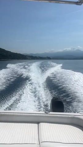 The video presents a view from the stern of a boat navigating calm, blue waters under a bright, clear blue sky. The main activity is the boat's steady movement, which creates a prominent, churning white wake trailing behind it.

The back of the boat is visible, featuring white, horizontally ribbed cushioned seating and a black outboard motor, where the brand name "MERCURY" is faintly discernable. White, empty fishing rod holders are mounted on the transom.

In the distance, a mountainous coastline stretches across the horizon, with some subtle structures visible along the shore. A few fluffy white clouds are scattered above the mountain peaks. The tranquil water beyond the boat's wake reflects the sky, appearing a deep, serene blue.

The scene depicts a sunny day, likely morning or early afternoon, with clear weather conditions ideal for boating. The setting is a picturesque coastal environment, specifically identified as Marina di Casal Velino, Italy, which aligns with the Mediterranean landscape visible. The continuous hum of the boat's engine is the only sound heard. No individuals are visible in the frame.