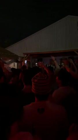 A crowd of people are gathered in an outdoor space at night. The background features a large, gray building with horizontal panels. Some people are dancing and raising their hands. Red lighting illuminates the scene.