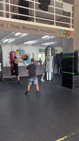 The video shows a boxing gym. In the foreground, a young person in athletic shorts and a t-shirt is holding a large orange ball. Behind them, a large black punching bag hangs from the ceiling. To the right, another person in red athletic shorts and a red top is throwing the orange ball. In the background, there are more punching bags, weight racks, and flags. The gym appears to be well-lit and has a dark, rubberized floor. The sign above reads "THE BOXING ACADEMY."