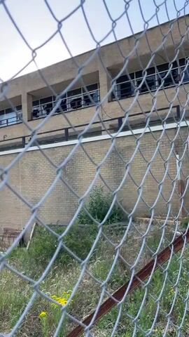 The video shows an abandoned building with broken windows, viewed through a chain-link fence. The building has a brick lower level and a tan upper level with a row of windows. Graffiti is visible on the upper level, including what appears to be "MARS & MONKEY" and "SOAP". The lower level has garage-style doors, some of which are boarded up. The area in front of the building appears to be a parking lot with debris and overgrown grass. The sky is partly cloudy. The video is shot from a low angle, looking up at the building.