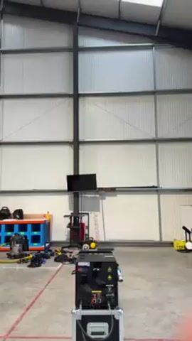 A device emitting two large bursts of fire is in the center of the screen. The device is black and on top of a gray case with handles. In the background is a large corrugated metal warehouse. To the left of the machine is a blue and orange storage cabinet. Behind the machine is a black tv mounted on a red rolling tool cart, a white board, and a variety of other tools and items. The camera is stationary and level with the horizon.