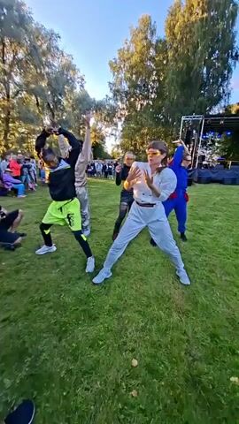 Three dancers perform an energetic routine on a grassy outdoor stage. The dancer on the left, a Black male, wears a black hoodie with lime green accents and matching lime green athletic pants. He executes a dynamic squatting pose with arms extended. The central dancer, a white female, is dressed in a light gray jumpsuit with puffy sleeves and sunglasses. She strikes a powerful, wide stance, one arm raised and bent. To the right, another dancer, possibly male, is clad in a blue jumpsuit with red accents. He mirrors the central dancer with a raised, bent arm. Behind them, a diverse crowd of onlookers watches from a distance. In the background, a stage with lighting equipment is visible, suggesting a performance or festival setting. The environment is a park with tall trees and a clear blue sky.
