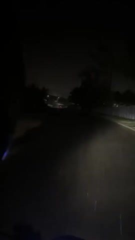 The video shows a dark road at night, with the headlights of a vehicle illuminating the path ahead. The road appears to be wet, suggesting it may have rained recently. Trees and houses are visible on either side of the road, silhouetted against the night sky. There are streetlights in the distance, casting a faint glow. The camera is positioned at a low angle, possibly from the perspective of someone riding a motorcycle or in a car. The video is short and shows no people or specific actions.