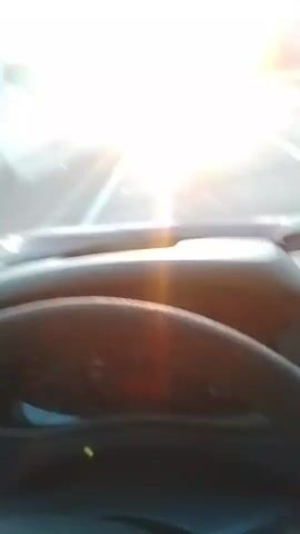 The video shows a first-person view from inside a vehicle traveling on a highway. The sun is setting or rising directly ahead, causing a strong glare that makes it difficult to see the road clearly. Other vehicles are visible in the distance on the highway. The steering wheel and dashboard of the vehicle are in the foreground. Trees and foliage line the sides of the highway.