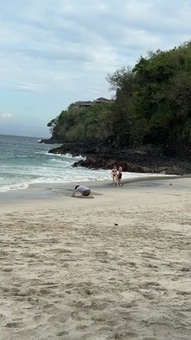 A wide shot captures a sandy beach with a person kneeling in the foreground, drawing in the sand. In the midground, a couple stands near the water's edge, looking towards the ocean. Behind them, waves crash against dark, rocky cliffs covered in lush green vegetation. A building with a red roof is visible on the hillside. The sky is overcast.