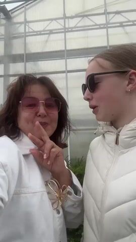 A medium shot shows two females standing close together, facing the camera. The female on the left, with dark hair and pink sunglasses, sticks her tongue out. She is wearing a white jacket with a gold butterfly pin. The female on the right, with blonde hair and black sunglasses, is smiling slightly. She is wearing a white puffer jacket. They appear to be in a greenhouse or similar structure with metal beams and translucent panels visible in the background. The lighting is overcast.