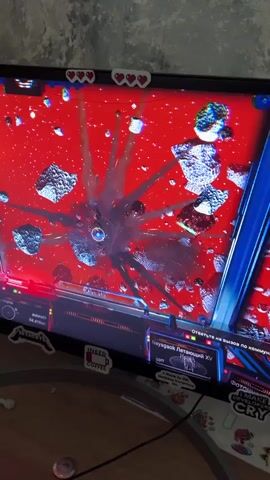 The video shows a child playing a video game on a monitor. The screen displays a space scene with a red background, asteroids, and a spaceship. The child is wearing a hooded sweatshirt and coughing. The monitor is surrounded by stickers. A keyboard and papers are on the desk next to the monitor.