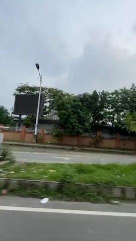 The video presents a street scene, captured from what appears to be a moving vehicle. The road stretches out ahead, marked with white lines, and is largely devoid of traffic. 

On the left side of the road, a low, reddish-brown wall is visible, partially obscured by greenery and trees. A yellow sign with text in a non-English script briefly appears. Further down the road, a yellow and red truck is parked on the roadside. Streetlights line the road.

To the right of the road, there is a concrete wall with a geometric pattern. The sky above is overcast with grey clouds.