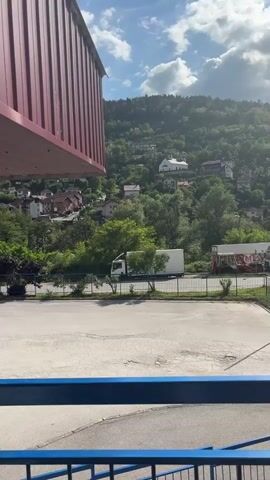 The video shows an outdoor area with a red roof and blue metal railings. A white bus with "TRANSERROM" written on its side is parked nearby. People are sitting and standing along the covered area, some waiting. The background features a hillside with buildings and greenery.