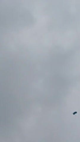 A wide shot captures a gray, cloudy sky. A small, dark blue object with a parachute deployed is visible in the lower right quadrant of the frame, descending. The object is the only distinguishable element against the overcast sky.