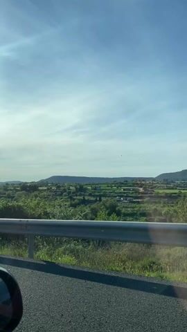 The video shows the landscape of Mallorca, Spain, from the perspective of a moving car. The view includes green fields, trees, and distant hills under a bright sky with light clouds. A metal guardrail is visible in the foreground, partially obscuring the view. A road sign is attached to the guardrail.