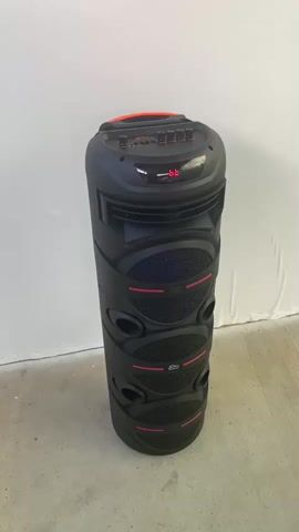 A tall, black cylindrical speaker with red accents stands against a white wall and a grey floor. The speaker has several circular speaker grilles stacked vertically and a control panel with buttons and a digital display at the top. The digital display shows "66". The camera moves slightly from left to right, showing the speaker from a slightly different angle.