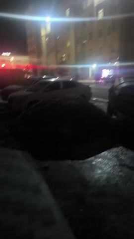 The video shows an urban nighttime scene.  Tall buildings with illuminated windows are in the background. Cars are parked along the street. Piles of dark material, possibly dirt or debris, are in the foreground. The camera moves slightly, eventually focusing on two men walking under a covered walkway. Light flares from streetlights or building lights are visible throughout the video.  The overall scene is somewhat dark and blurred.
