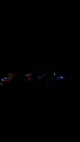 A dark video of a group of people silhouetted against dimly lit stage lights. The lighting is primarily red, purple, and green, casting colorful glows. Several individuals appear to be on a stage or elevated platform, possibly performing or setting up. There's a sense of a concert or event in progress, with a crowd of people visible in the foreground, also in silhouette against the stage lights. The overall scene is very dark, making detailed identification of individuals or objects difficult.