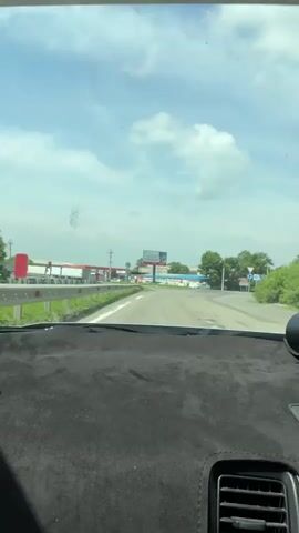 The video shows a view from the inside of a car looking out through the windshield. The car is on a road, and in the distance, there is a gas station with several pumps and buildings. A large billboard is visible above the gas station, featuring text and images. The sky is blue with white clouds. The road curves to the right in the distance. The dashboard of the car is partially visible in the foreground, covered with black material.