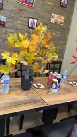 The camera pans across a wooden table in a casual dining area.  On the table are two artificial trees with fall foliage, a stack of paper cups, several cans of soda, two bottles of water, and two small plates, each containing a single brownie. A wall behind the table is decorated with colorful graffiti and various framed pictures. One wall is covered in a stone-like material.  The lighting is bright.