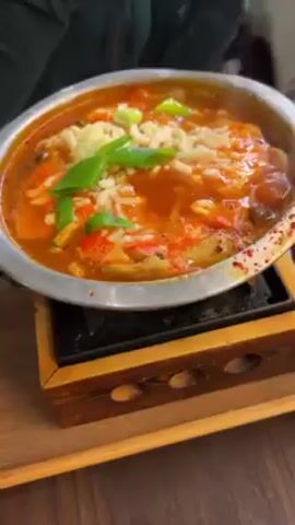 A close-up video shows a hot, bubbling dish, likely a stew or soup, in a silver bowl. The dish appears to be tomato-based, containing what seems like sliced vegetables (possibly bell peppers), mushrooms, and noodles. It's topped with shredded cheese and sliced green onions for garnish. The bowl is sitting on a black hot plate, which is in turn resting on a wooden tray with cut-out designs. The video captures the steaming and bubbling of the dish as it cooks. In the beginning, a person is partially visible in the left side of the frame. Another person, wearing a green shirt, reaches in to adjust the bowl.