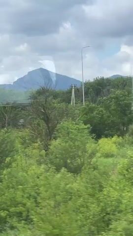 The video captures a view from inside a vehicle, looking out at a landscape dominated by lush green trees and vegetation. In the distance, mountains are visible under a cloudy sky. The camera appears to be moving, causing a slight blur to the scenery. Reflections on the vehicle's window are also noticeable, showing a distorted image of the interior and the outside world. The lighting suggests it is daytime, possibly overcast. There are no people or specific objects clearly identifiable within the frame other than the natural environment and elements of the vehicle.