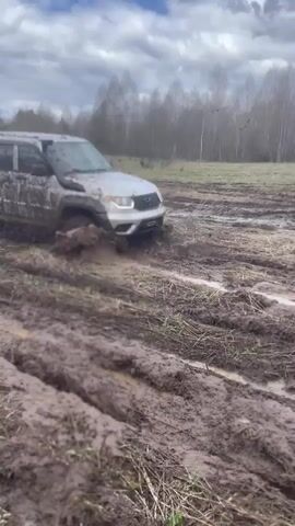 A silver SUV covered in mud navigates a muddy field. The SUV is equipped with an air intake snorkel. A person in a red jacket and tan hat kneels in the foreground, taking a picture of the SUV with a phone. The sky is overcast, and trees border the field in the background. The camera focuses on the SUV as it churns through the mud.