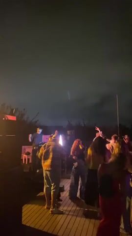 A group of people are dancing outdoors at night. The ground is made of wood planks. There are lights, including what appears to be a spotlight or a flame in the center of the frame, illuminating the scene. Several people are visible from behind, with their backs to the camera, as they dance. The sky is dark, indicating it is nighttime.