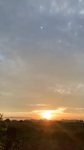 This video shows a sunset over a rural landscape. The sun is low on the horizon, casting a warm, golden light across the scene. The sky is filled with clouds, which are illuminated by the setting sun. In the foreground, there is a field of grass and some trees. In the background, there are some hills and mountains. The overall mood of the video is peaceful and serene.