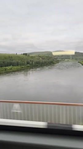 The video shows a view from a moving vehicle, likely a train, looking out over a wide river. The river is calm with some ripples on the surface. The banks of the river are lined with green trees and rolling hills in the background. The sky is overcast with clouds. In the distance, on top of a hill, there is a bright, yellowish rectangular object that is partially obscured by clouds. The foreground shows a portion of the vehicle's interior, including a railing with vertical white and black stripes. The video is a brief, static shot of the landscape.