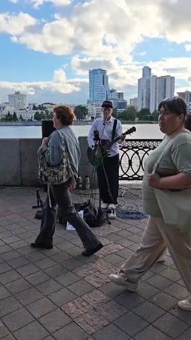 The video shows a street musician playing an acoustic guitar in an outdoor urban setting. The musician is standing next to a speaker and some music equipment. They are wearing a white shirt and dark pants. In the foreground, several people are walking past, some are in focus, others are blurred. The background features a body of water, a decorative fence, and a cityscape with buildings and a blue sky with clouds. The video appears to be filmed from a moving perspective, following the people walking.