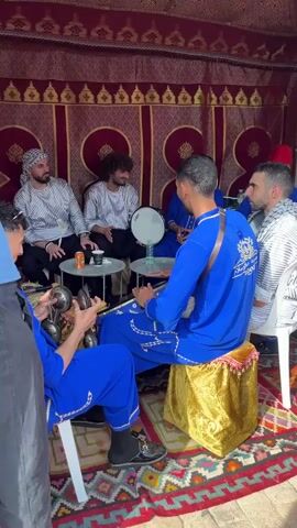 Inside a tent decorated with red and gold tapestries, a group of men are making music. One man wears a blue robe embroidered with gold, and sits on a gold-colored cushion. He plays a large drum. Another man in a blue robe plays cymbals. One man plays a tambourine, and three others sit on chairs around a small table. They wear striped shirts and drink from cans and cups. Someone in a blue shirt stands to the left, partially cut off by the frame.