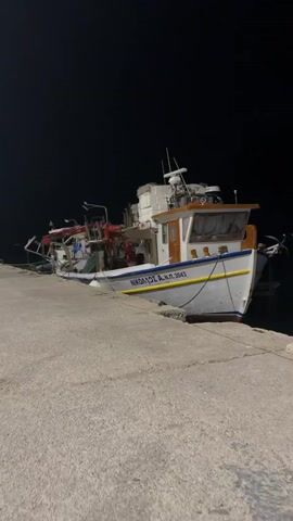 A fishing boat named NIKOLAOS A.N.P. 3043 is docked at a pier at night. The boat is white with a blue and orange stripe and has a cabin structure at the rear. Fishing equipment is visible on the deck. The pier is made of concrete. The background is dark, indicating it is nighttime. The camera is moving slightly, causing some blur.