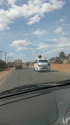 The video shows a drive along a paved road through a semi-urban environment under a partly cloudy sky. Several large trucks are driving in the same direction, and there are numerous people walking and congregating along the roadside. Power lines are visible along the road. The environment appears dry, with dusty areas off the pavement. A motorcycle with two riders passes by. The view is from inside a car, focusing on the road ahead.