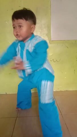 A young boy wearing a blue shirt and pants is on the floor. He is in a push-up position and appears to be struggling or falling. His body is blurred due to movement. He is on a green floor with tile flooring visible in the background.