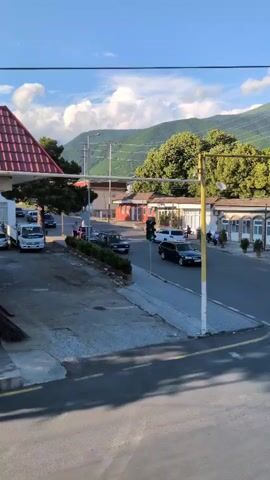 This media file captures a street scene in Şəki, Azerbaijan, during what appears to be the late afternoon or early evening, given the warm sunlight and long shadows. The weather is pleasant, with a clear blue sky dotted with fluffy white clouds and lush green mountains forming the backdrop.

The scene is set on a moderately busy street. Several vehicles are visible, including a white truck, a dark sedan, and what looks like an older model dark-colored car. A woman is seen walking across a crosswalk. A traffic light is illuminated green, indicating traffic flow. People are casually gathered outside some of the buildings lining the street, suggesting a relaxed atmosphere.

The architecture includes buildings with red-tiled roofs and some with stone facades. There are also power lines and utility poles, characteristic of a town or city. The overall impression is one of a peaceful, everyday moment in a town nestled among scenic mountains. No discernable text is clearly visible within the frame.