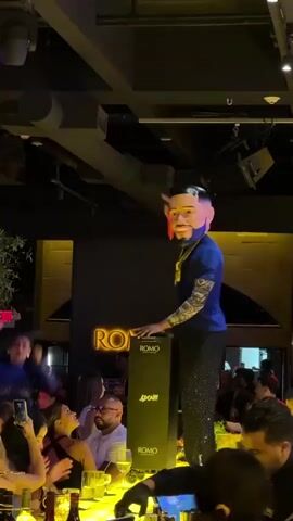The video shows a person wearing a large, exaggerated mask of a face. The person is standing on a raised platform or stage, dressed in a blue t-shirt, black pants with sparkling accents, and a gold chain. They are operating a DJ setup which includes a black speaker-like console with the word "ROMO" visible multiple times. The person is gesturing with their hands and appears to be interacting with the music or the crowd. Around the stage, a crowd of people are visible, some are seated at tables with drinks, others are standing and appear to be watching the performance. The environment is dimly lit with colored stage lights, predominantly green and yellow. A neon sign with the word "ROMO" is visible in the background. The overall atmosphere seems to be that of a nightclub or a party.