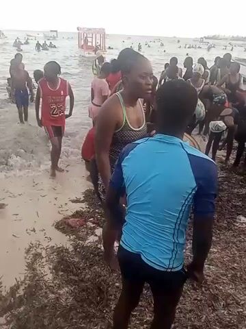 The video shows a crowded beach with many people, mostly children, standing in shallow water near the shore. Several boats or floating structures are visible in the distance.  A man carries a large black tire tube, initially around his neck, then transferring it to his arm, and eventually handing it to a woman. Some seaweed is scattered on the sand near the waterline. The overall scene appears lively and recreational.