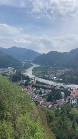 A drone shot pans over a valley with a river winding through it. A town with red-tiled roofs sits on the banks of the river, nestled amongst rolling green hills. The sky is partly cloudy, with patches of blue peeking through. In the foreground, the lush green foliage of trees frames the view. The camera moves slightly to the right, revealing more of the town and the surrounding mountains. The scene is peaceful and picturesque.