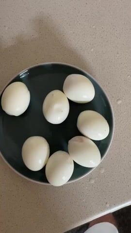 This media file features a close-up, top-down view of seven peeled hard-boiled eggs arranged on a dark green plate. The plate is placed on a speckled, light-colored countertop, which appears to be a kitchen surface. A few water droplets are visible on the countertop around the plate, suggesting the eggs may have recently been cooked or rinsed.

The eggs are uniformly white and appear to be fully cooked. There are no visible yolks or any other ingredients associated with preparing dishes like deviled eggs. The lighting in the scene is bright, indicating it is likely daytime, and there are no discernable external light sources like windows or lamps casting specific shadows, suggesting ambient indoor lighting.

The overall impression is one of food preparation or presentation. The arrangement of the eggs is casual, with some touching and others slightly spaced apart. The focus is entirely on the eggs and the plate, with no other objects or people clearly visible in the frame, except for a small portion of what looks like a white sock or shoe in the bottom right corner, hinting at a person's presence in the vicinity.

There is no visible text in this media file. The context of Reading, United Kingdom, does not provide any specific visual cues within the image itself.