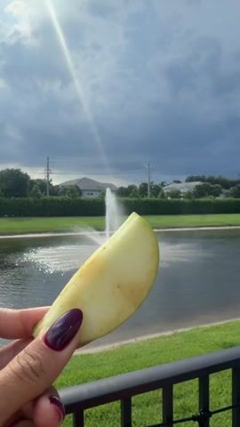 A hand with dark purple fingernails holds a slice of green apple in the foreground. Behind the apple, a water fountain sprays water into a pond. Trees and houses are visible in the background, under a cloudy sky with a sunbeam. The person takes a bite of the apple.