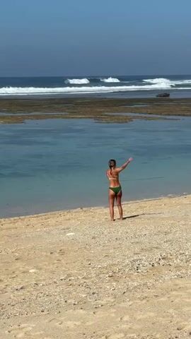 A woman in a green bikini stands on a sandy beach facing the ocean. She has her arms extended to her sides. In the background, the ocean has waves breaking on a reef. The sky is clear and blue.