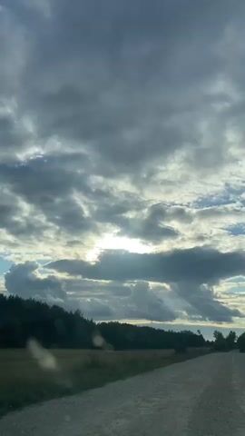 The video shows a view from a vehicle moving along a dirt road. The sky is filled with dramatic clouds, with rays of sunlight breaking through. To the left of the road is a field, and beyond that, a dense line of trees. The road curves slightly to the right as it extends into the distance. There are no people or other vehicles visible. The overall impression is of a natural landscape under a dynamic sky.