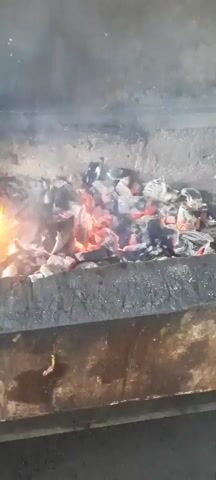 A close-up shot shows a pile of burning charcoal in a grill or fire pit. The charcoal glows with red and orange embers, and wisps of smoke rise from the fire. The surrounding area appears to be made of concrete or stone, with some dark smudges or stains. The lighting is dim, highlighting the intense heat and light of the fire.